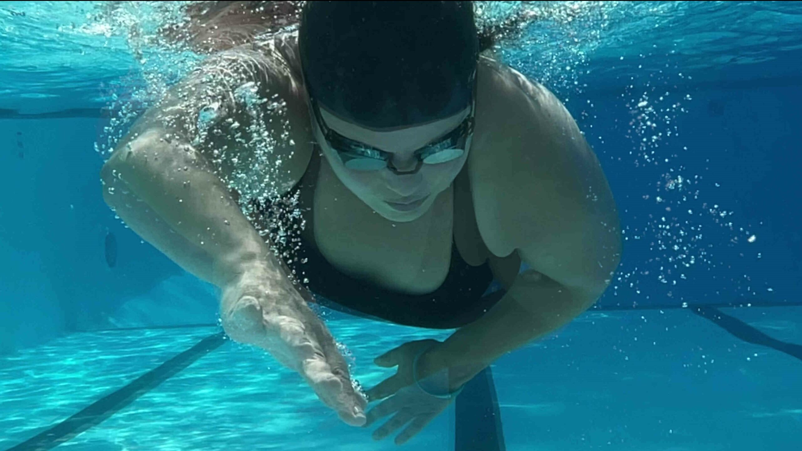 underwater shot of a fair-skinned woman in a black bathing suit and swim cap wearing goggles as she swims in a pool